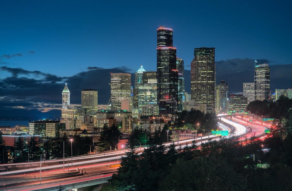 The Seattle skyline at dusk, seen from the Jose Rizal Bridge with Interstate 5 and its car light trails in the foreground.